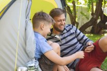 Homme caucasien passant du temps avec son fils ensemble, campant dans le jardin, allongé dans une tente à l'aide d'un smartphone, souriant. Distance sociale pendant le confinement en quarantaine du coronavirus Covid 19. — Photo de stock