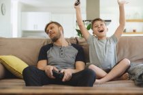 Caucasian man at home with his son together, sitting on sofa in living room, playing video games, boy cheering victory with arms up. Social distancing during Covid 19 Coronavirus quarantine lockdown. — Stock Photo