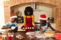 African american mother wearing apron holding baked cookies and daughter and son wearing santa hat baking in the kitchen at home. christmas festivity tradition celebration concept — Stock Photo