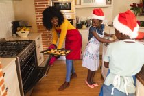 Une mère afro-américaine portant un tablier contenant des biscuits cuits et une fille et un fils portant un chapeau de Père Noël cuisinant dans la cuisine à la maison. Noël fête tradition célébration concept — Photo de stock