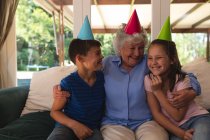 Старшая кавказская женщина проводит время дома, празднуя день рождения с внуками, надевая праздничные шляпы и улыбаясь. Качество времени вместе в коронавирусном ковиде 19 блокировка карантина. — стоковое фото