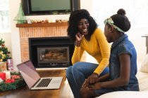 Madre e hija afroamericanas sonriendo mientras tienen un video chat en el portátil en la sala de estar en casa. distanciamiento social durante el bloqueo de cuarentena por coronavirus covid 19. - foto de stock