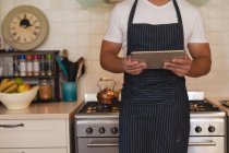 Caucasian man standing in a kitchen and wearing apron, using his tablet. self isolation at home during coronavirus covid 19 quarantine lockdown. — Stock Photo