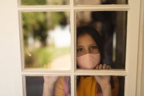Portrait of caucasian girl wearing face mask looking at camera through window. protection and self isolation during covid 19 coronavirus pandemic lockdown. — Stock Photo