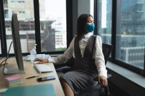 Asiatin mit Gesichtsmaske sitzt auf ihrem Schreibtisch und blickt aus dem Fenster auf modernes Büro. Soziale Distanzierung von Quarantäne während der Coronavirus-Pandemie — Stockfoto