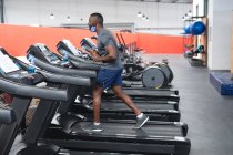Fit african american man wearing face mask running on treadmill doing cardio workout in the gym. social distancing quarantine lockdown during coronavirus pandemic — Stock Photo