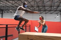 Fit african american man wearing face mask jumping on wooden plyo box in the gym  while  caucasian female fitness trainer holding stopwatch and clipboard. social distancing quarantine lockdown during coronavirus pandemic — Stock Photo