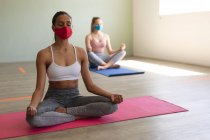 Two fit caucasian women wearing face masks practicing yoga while sitting on yoga mats in the gym. social distancing quarantine lockdown during coronavirus pandemic — Stock Photo