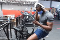 Жирний афроамериканець, одягнений в маску обличчя і навушники за допомогою смартфона, сидячи на стаціонарному велосипеді в спортзалі. Відстань до карантину під час пандемії коронавірусу — стокове фото