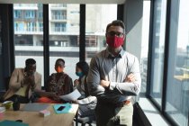 Retrato de hombre caucásico con máscara facial de pie con los brazos cruzados en la oficina moderna. distanciamiento social bloqueo de cuarentena durante la pandemia de coronavirus - foto de stock