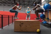 Fit african american man and fit  caucasian woman wearing face masks jumping on wooden plyo box in the gym while  caucasian female fitness trainer holding stopwatch and clipboard. social distancing quarantine lockdown during coronavirus pandemic — Stock Photo