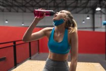 Fit  caucasian woman wearing face mask around her neck drinking water while sitting on wooden plyo box in the gym. social distancing quarantine lockdown during coronavirus pandemic — Stock Photo