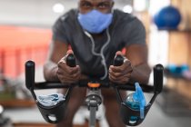 Портрет сильного африканского американца в маске для лица и наушниках, упражняющихся на стационарном велосипеде в тренажерном зале. социальная изоляция от карантина во время пандемии коронавируса — стоковое фото