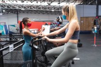 Fit caucasian woman wearing face mask exercising on stationary bike while caucasian female fitness coach taking notes on clipboard  in the gym. social distancing quarantine lockdown during coronavirus pandemic — Stock Photo