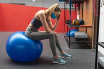 Stressed fit caucasian woman wearing face mask sitting on exercise ball in the gym. social distancing quarantine lockdown during coronavirus pandemic — Stock Photo