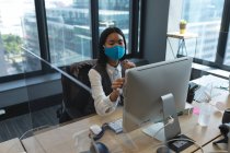 Азійка, одягнена в маску обличчя, за допомогою комп'ютера сидить на столі в сучасному офісі. Відстань до карантину під час пандемії коронавірусу — стокове фото