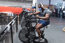 Ajuste hombre afroamericano con máscara facial y auriculares haciendo ejercicio en bicicleta estacionaria en el gimnasio. distanciamiento social bloqueo de cuarentena durante la pandemia de coronavirus - foto de stock