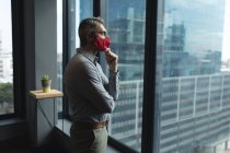 Homem caucasiano cuidadoso usando máscara facial olhando pela janela no escritório moderno. bloqueio de quarentena por distanciamento social durante a pandemia do coronavírus — Fotografia de Stock