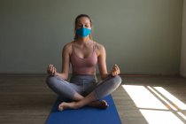 Впишеться біла жінка в маску для обличчя, що практикує йогу, сидячи на килимку для йоги у спортзалі. карантин соціального дистанціювання під час пандемії коронавірусу — стокове фото