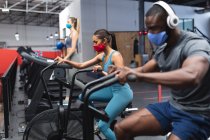 Fit african american man and fit caucasian woman wearing face masks exercising on stationary bike in the gym. social distancing quarantine lockdown during coronavirus pandemic — Stock Photo