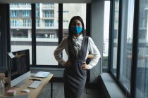 Porträt einer asiatischen Frau mit Gesichtsmaske, die mit den Händen auf den Hüften im modernen Büro steht. Soziale Distanzierung von Quarantäne während der Coronavirus-Pandemie — Stockfoto