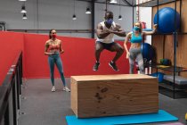 Fit african american man wearing face mask jumping on wooden plyo box in the gym while caucasian female fitness trainer holding stopwatch. social distancing quarantine lockdown during coronavirus pandemic — Stock Photo