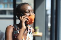 Mulher afro-americana usando máscara facial falando no smartphone no escritório moderno. bloqueio de quarentena por distanciamento social durante a pandemia do coronavírus — Fotografia de Stock