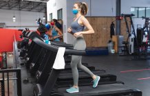 Fit caucasian woman wearing face mask running on treadmill doing cardio workout in the gym. social distancing quarantine lockdown during coronavirus pandemic — Stock Photo