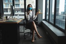 Азиатка в маске сидит на столе в современном офисе. социальная изоляция от карантина во время пандемии коронавируса — стоковое фото