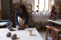 Mixed race female potter in face mask working in pottery studio. wearing apron, working at a working table with her friend in the back. small creative business during covid 19 coronavirus pandemic. — Stock Photo