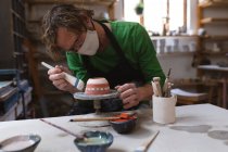 Ceramista maschio caucasico in maschera facciale che lavora in studio di ceramica. indossando grembiule, lavorando alla ruota di un vasaio, dipingendo ciotola. piccola attività creativa durante covid 19 coronavirus pandemia. — Foto stock