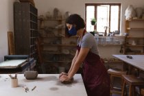 Ceramista caucasica in maschera facciale che lavora in studio di ceramica. indossando grembiule, lavorando ad un tavolo di lavoro. piccola attività creativa durante covid 19 coronavirus pandemia. — Foto stock