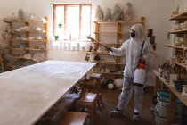 Белый мужчина-уборщик в защитной одежде работает в мастерской керамики. Дезинфекция всего места. малый творческий бизнес во время пандемии коронавируса ковида 19. — стоковое фото