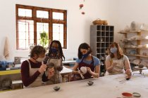 Grupo multiétnico de alfareros con máscaras faciales que trabajan en un estudio de cerámica. Usando delantales, pintando platos. pequeña empresa creativa durante la pandemia de coronavirus covid 19. - foto de stock
