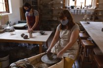 Two caucasian female potters in face masks working in pottery studio. wearing apron, working at a potters wheel and working table. small creative business during covid 19 coronavirus pandemic. — Stock Photo
