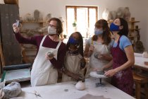 Multi-ethnic group of potters in face masks working in pottery studio. wearing aprons, painting plates, taking a selfie together. small creative business during covid 19 coronavirus pandemic. — Stock Photo