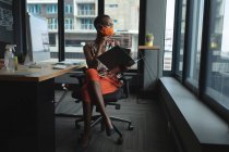 Uma afro-americana a usar máscara facial a trabalhar no escritório. sentado em documentos de leitura de mesa. higiene e distanciamento social no local de trabalho durante coronavírus covid 19 pandemia. — Fotografia de Stock
