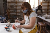 Кавказька жінка-гончар в масці обличчя працює в гончарній студії. одягнений в фартух, працюючи за робочим столом, малював тарілку. маленьке творче підприємство під час 19-ї коронавірусної пандемії. — стокове фото
