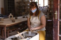 Caucasian female potter in face mask working in pottery studio. wearing apron, working at a potters wheel. small creative business during covid 19 coronavirus pandemic. — Stock Photo