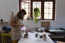 Potier femme caucasienne en masque facial travaillant dans un atelier de poterie. portant un tablier, travaillant à une table de travail, peignant un bol. petite entreprise créative pendant la pandémie de coronavirus covid 19. — Photo de stock