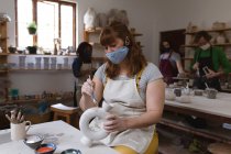 Кавказька жінка-гончар в масці обличчя працює в гончарній студії. в фартусі, за робочим столом зі своїми друзями на задньому плані. маленьке творче підприємство під час 19-ї коронавірусної пандемії. — стокове фото