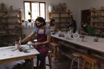 Caucasian female potter in face mask working in pottery studio. wearing apron, working at a working table with her friends in the back. small creative business during covid 19 coronavirus pandemic. — Stock Photo