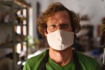 Retrato de un hombre caucásico usando mascarilla en un estudio de cerámica. pequeña empresa creativa durante la pandemia de coronavirus covid 19. - foto de stock