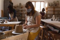 Caucasian female potter in face mask working in pottery studio. wearing apron, working at a potters wheel with two of her friends behind. small creative business during covid 19 coronavirus pandemic — Stock Photo