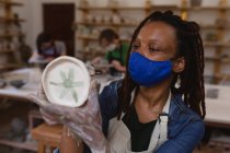 Ceramista di razza mista in maschera facciale che lavora in studio di ceramica. indossando un grembiule, lavorando ad un tavolo di lavoro, dipingendo un piatto. piccola attività creativa durante covid 19 coronavirus pandemia. — Foto stock