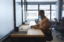 Homme afro-américain portant un masque facial à l'aide d'un ordinateur portable tout en étant assis sur son bureau au bureau moderne. isolement social mise en quarantaine pendant une pandémie de coronavirus — Photo de stock