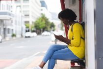 Africano mulher americana usando smartphone em uma rua. sentado em um banco e ouvir música com fones de ouvido em. fora e sobre na cidade durante covid 19 coronavirus pandemia. — Fotografia de Stock