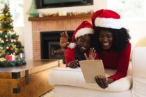 African american mother and daughter having video chat on tablet wearing santa hats during christmas at home social distancing during covid 19 coronavirus quarantine lockdown. — Stock Photo