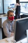 Белый мужчина в маске смотрит на экран компьютера в офисе. социальная изоляция от карантина во время пандемии коронавируса. — стоковое фото