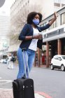Mujer afroamericana con mascarilla en la calle sosteniendo una taza de café y levantando la mano. fuera de la ciudad durante la pandemia de coronavirus covid 19. - foto de stock